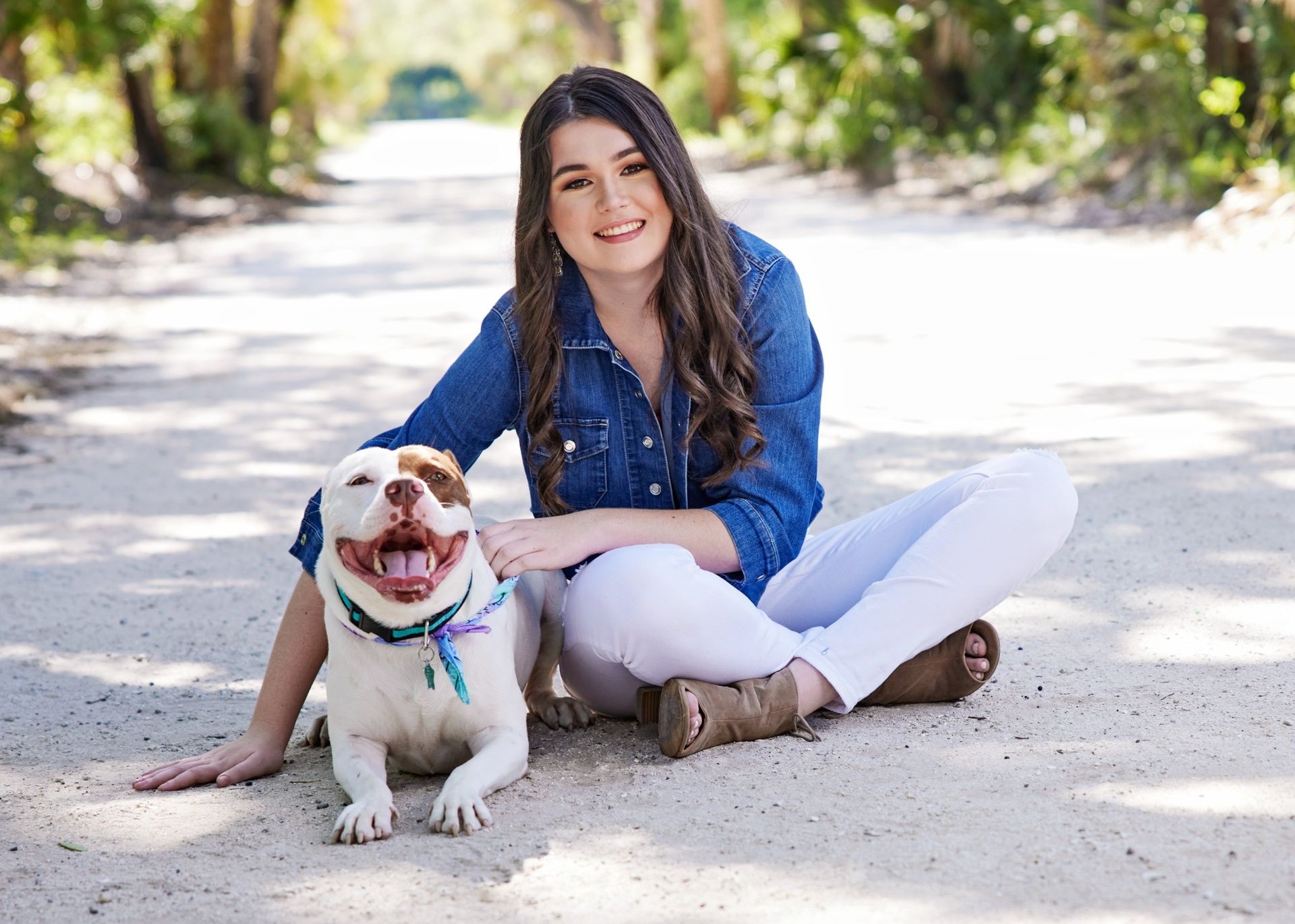 Professional outdoor portrait of a young woman with long brown hair, sitting on a sunlit path in a denim shirt and white pants, smiling beside a happy white and brown dog wearing a turquoise collar, photographed in Melbourne, Florida.
