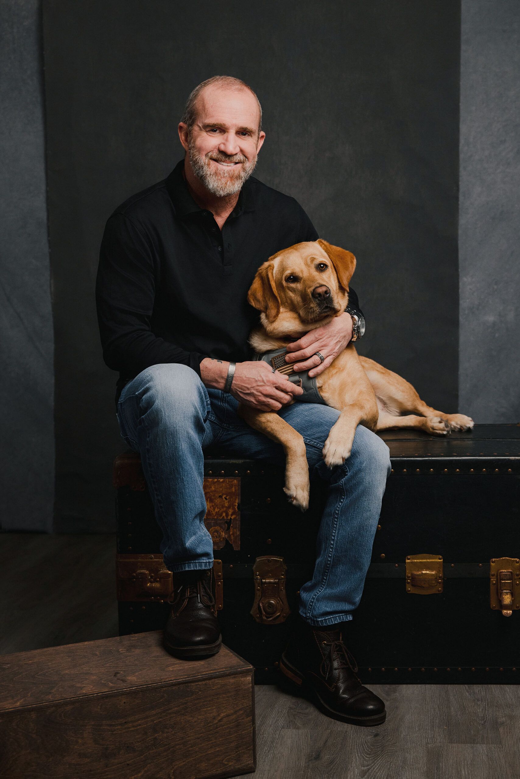 Pet and owner portrait featuring a smiling man holding his yellow Labrador Retriever while seated on a vintage trunk against a dark studio background.