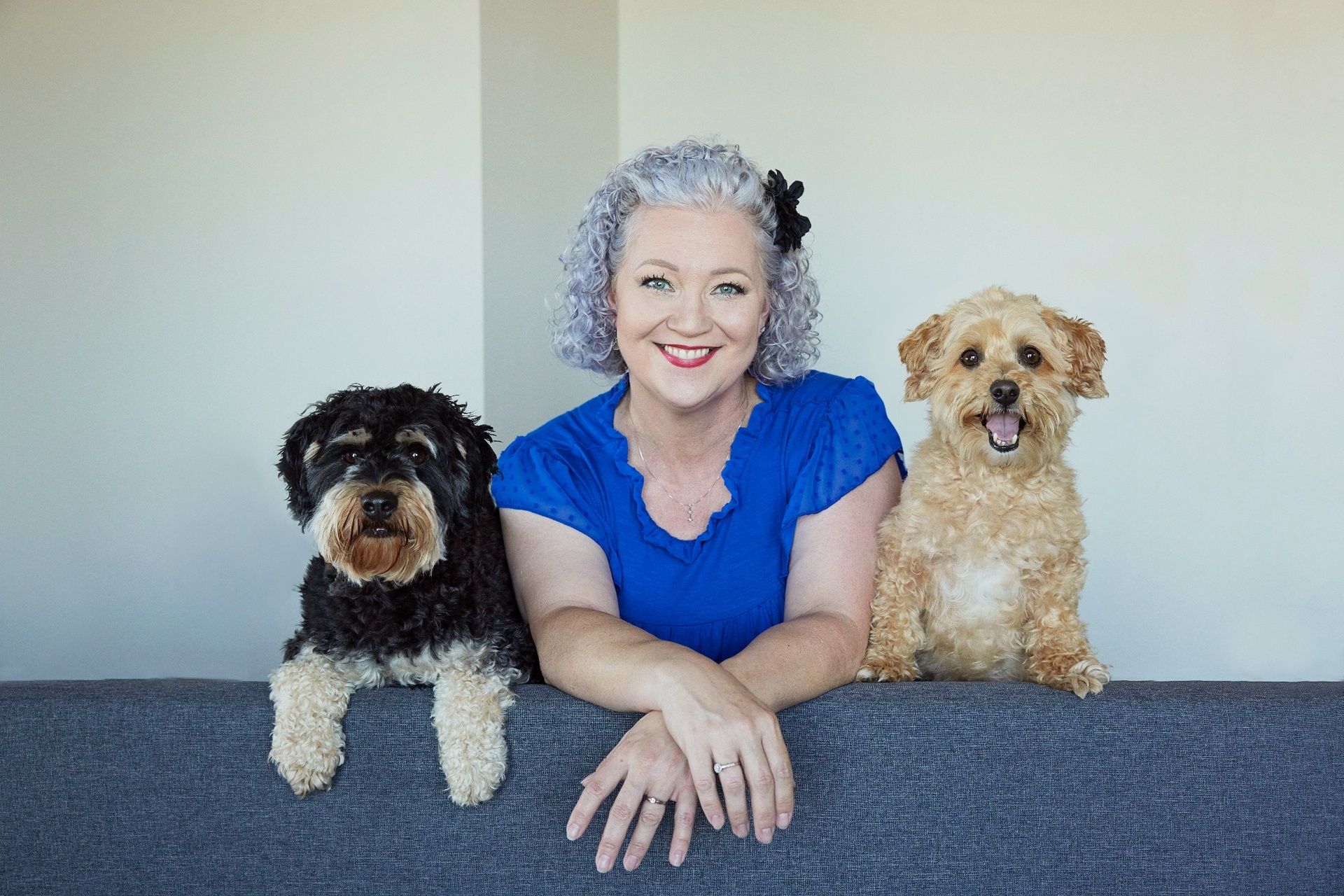 Professional personal branding portrait of a smiling woman with curly gray hair wearing a bright blue blouse, seated on a gray couch between two small dogs, one black-and-tan and one tan, photographed near Portland Oregon