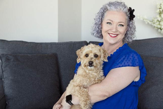 Woman with curly gray hair wearing a blue blouse, smiling while holding her small tan dog on a sofa during a pet portrait session.