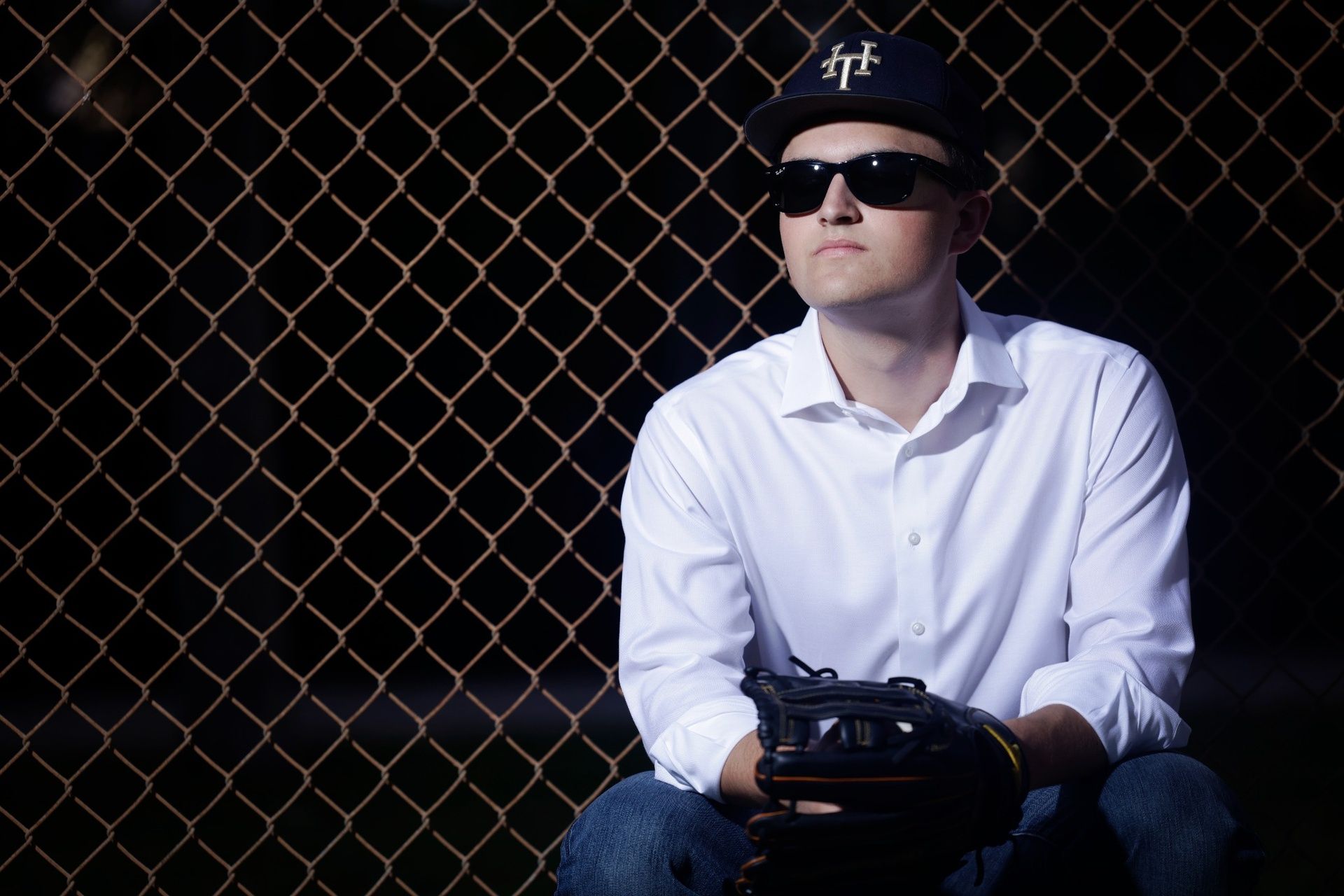 Professional portrait of a young man wearing sunglasses and a baseball cap, seated in front of a chain-link fence while holding a glove, dressed in a white shirt and jeans, photographed in Melbourne, Florida.”