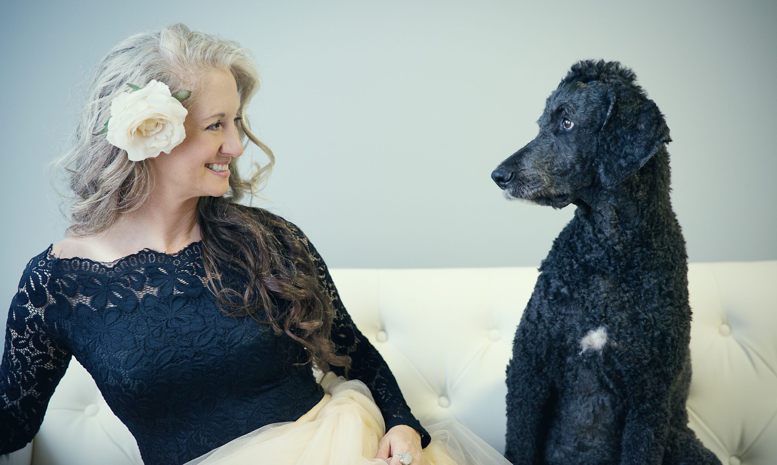 Fine art pet portrait of a woman in a black lace dress smiling at her black Poodle while seated together on a white sofa.