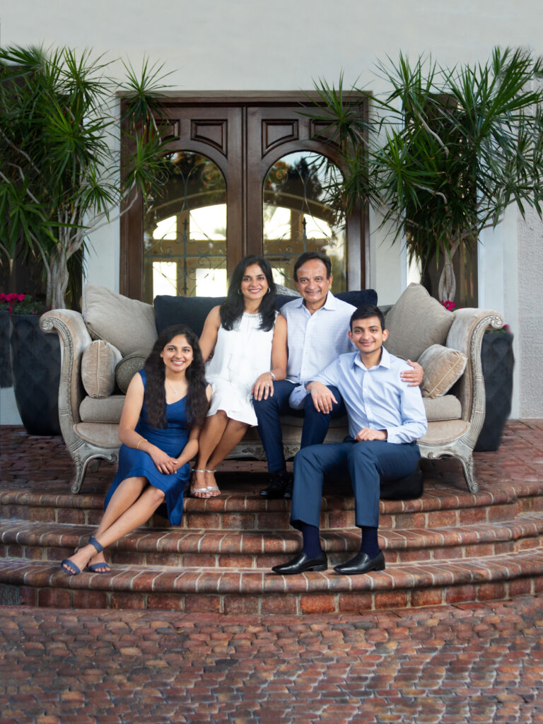 FAmily sitting on beige velvet couch in front of their home in front of their front doors. Dressed in navy, blues and white