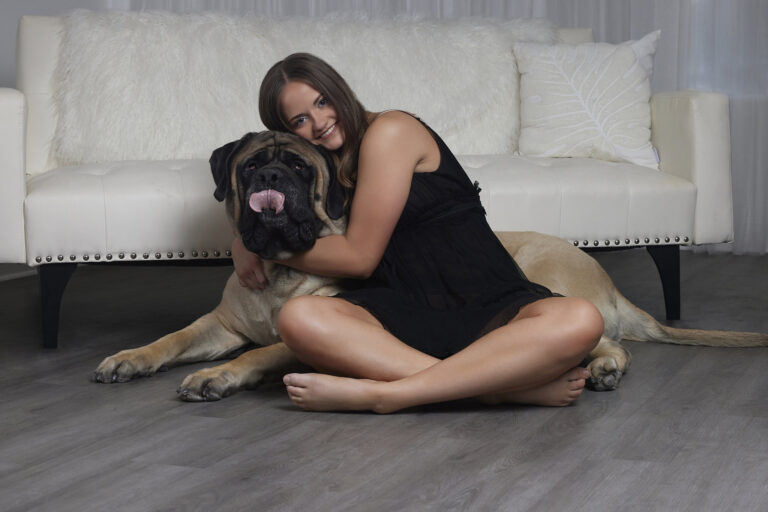 A woman sitting on the floor in a black dress, smiling and hugging a large English Mastiff dog with its tongue out. They are posed in front of a white couch in a modern, cozy living space