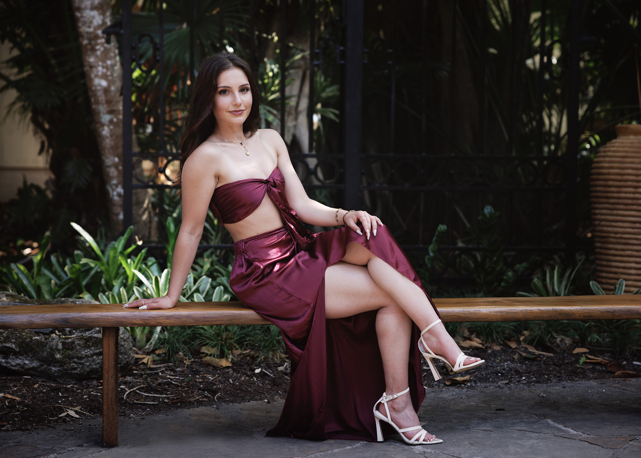 A young woman in a stunning burgundy dress sits on a wooden bench in an elegant pose. She is set against a lush outdoor background, exuding grace and confidence in her glamorous attire. She is a high School Senior