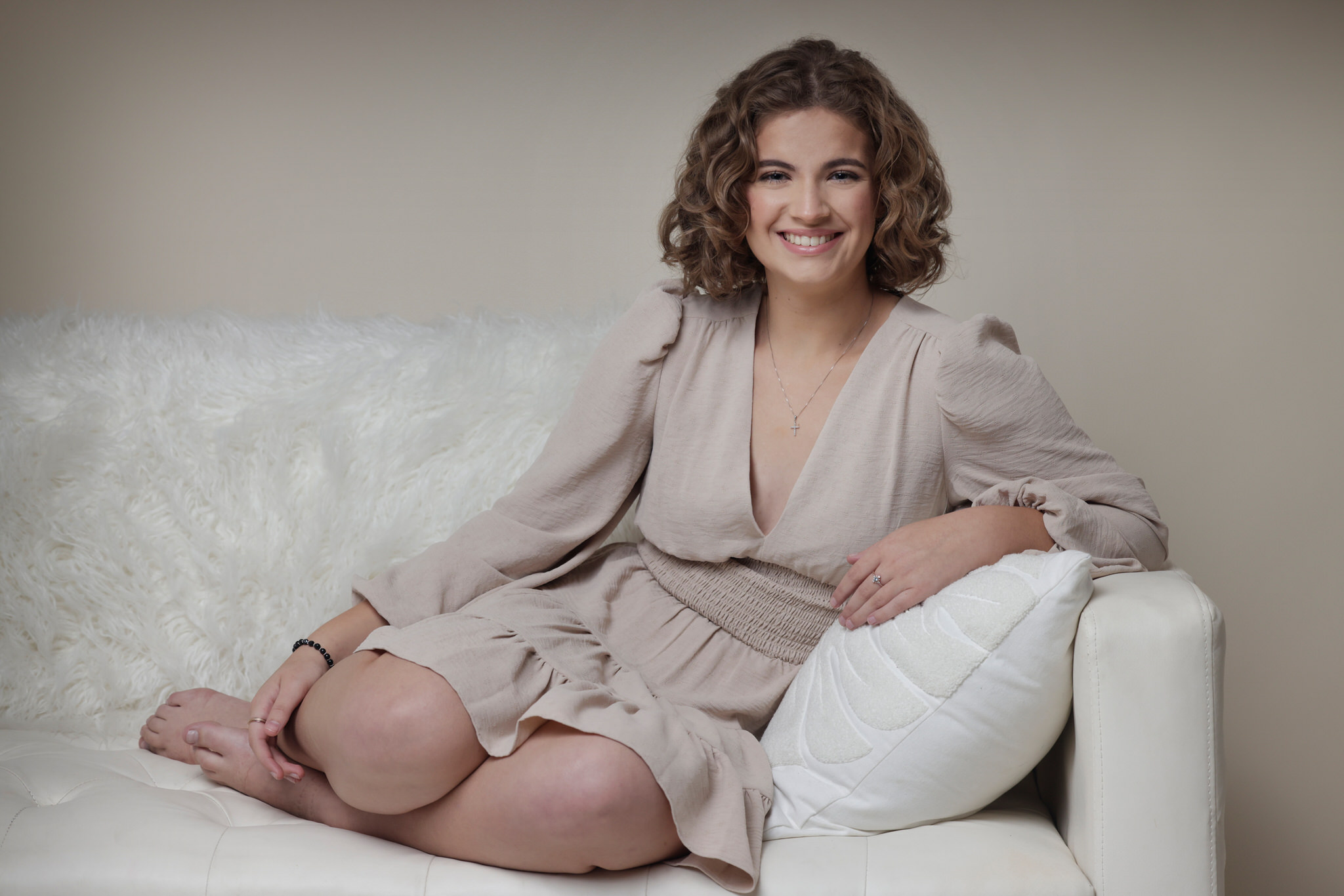 High school age caucasion girl with curly light brown hair sitting on a white couch leaning in the arm with her elbow. her feet are tucked under her